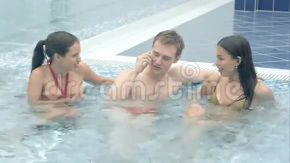 两个漂亮的女人嘲笑那个在游泳池里放松的年轻人接电话视频的预览图