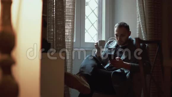 年轻人坐在客厅的扶手椅上使用智能手机有魅力的男性浏览互联网喝咖啡视频的预览图