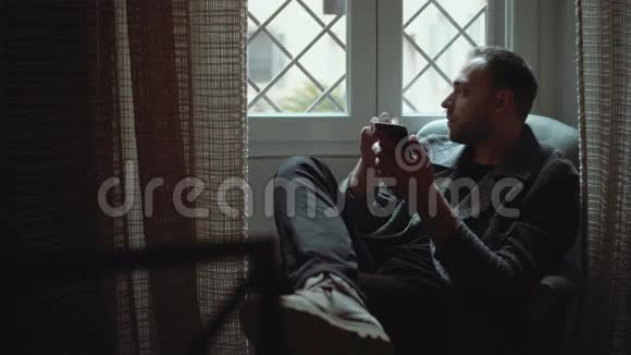 时髦的男人坐在扶手椅上看着窗户年轻迷人的男性在客厅里使用智能手机视频的预览图