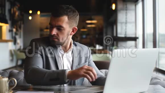 一个穿着灰色时髦西装的年轻商人在靠近窗户的一家咖啡馆的笔记本电脑上工作视频的预览图