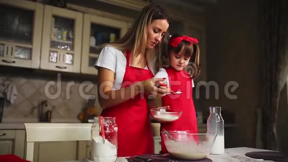 漂亮的年轻母亲帮她的小女儿用红色围裙做蛋糕倒牛奶搅拌面糊视频的预览图