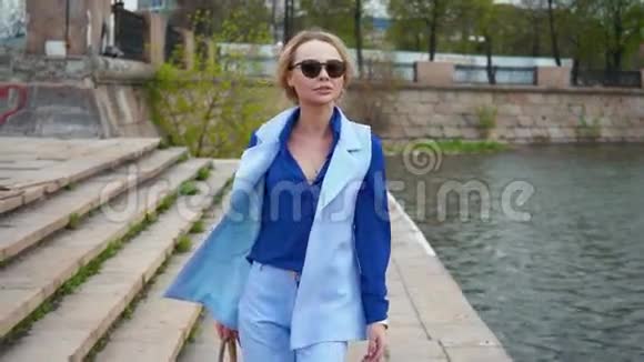 美丽的金发商务女性戴着太阳镜穿着蓝色西装在河边等着在城里见面放松一下视频的预览图