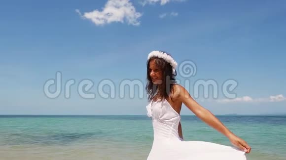 美丽的年轻女子穿着白色连衣裙和花圈光着脚在海岸边奔跑享受着旋转的乐趣视频的预览图