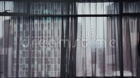 年轻的女人跑着打开大窗户上的窗帘看着她在城市建筑上的公寓慢动作视频的预览图