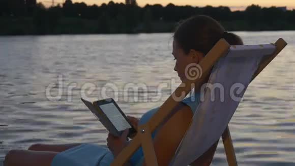 女孩在日落时分在水边的日光浴下阅读电子书视频的预览图