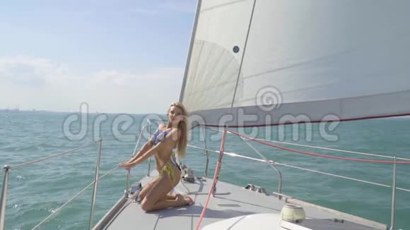 一个穿着蓝色比基尼的苗条金发女郎跪在游艇的船头上慢动作视频的预览图