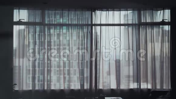 年轻女子的剪影在大窗户上拉开窗帘望着城市建筑上的公寓视频的预览图