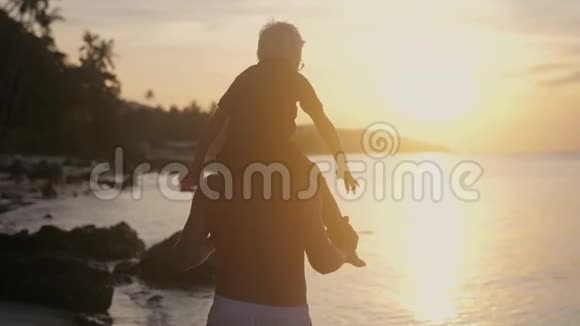 快乐的父亲和儿子戴着眼镜在热带海滩上玩耍男孩举起双手模仿美妙的日落飞行视频的预览图