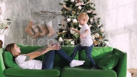 妈妈带着一个孩子在圣诞树背景的沙发上玩耍圣诞节室内视频的预览图
