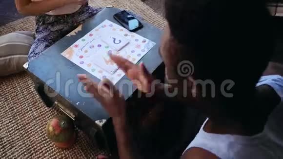 男人掷骰子坐在女人面前穿着睡衣的多种族夫妇在地板上玩棋盘游戏视频的预览图