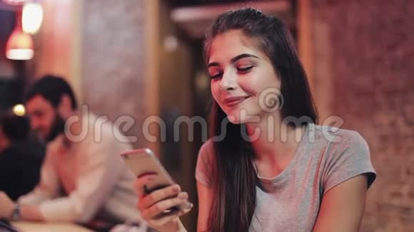 年轻迷人的女人美丽的微笑是使用智能手机在晚上坐在酒吧或餐馆里的女孩视频的预览图