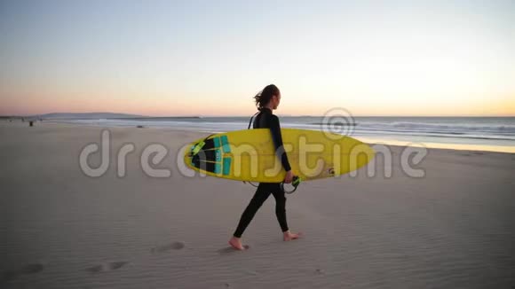 小女孩拿着冲浪板在海滩上带着海浪走向海洋的女人美丽的日落风吹沙子视频的预览图