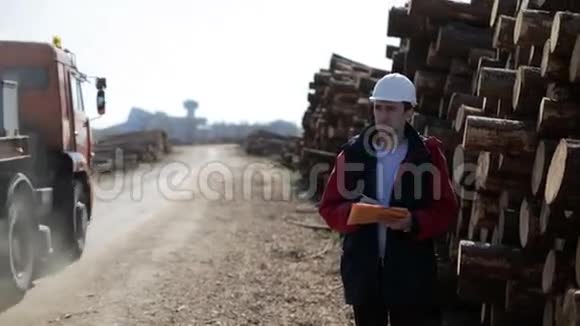 戴白色头盔的建筑工人站在原木旁边的道路上并监测货物运输视频的预览图