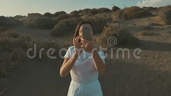 穿着白色连衣裙的女孩在日落时分在沙漠里打电话拍照温暖的夏日傍晚沙漠慢动作视频的预览图