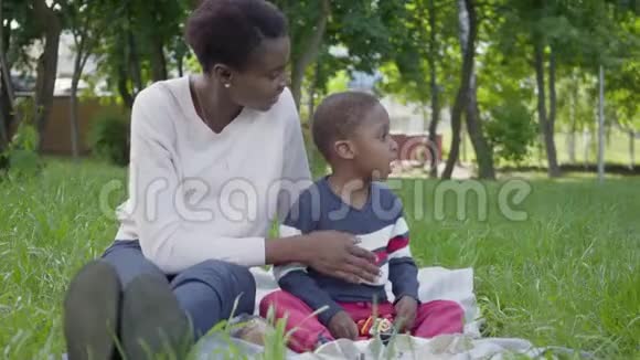 肖像可爱的非裔美国妇女坐在毯子上她的小儿子在公园里玩玩具视频的预览图