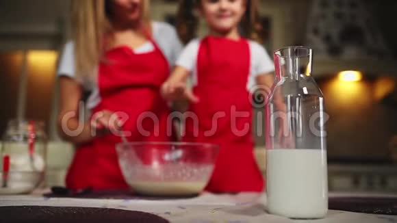 漂亮的年轻母亲帮她的小女儿用红色围裙做蛋糕倒牛奶搅拌面糊视频的预览图