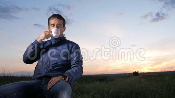 日落时一个年轻人从杯子里喝一杯热饮料后面是美丽的天空视频的预览图