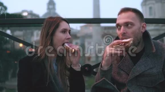 年轻时尚的夫妇晚上坐在市中心吃三明治一起往旁边看视频的预览图
