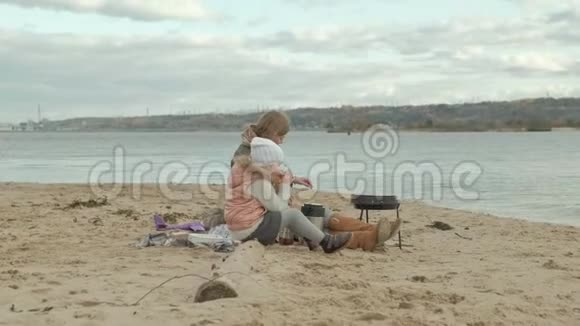 年轻的女人穿着一件外套有一个卷发的女孩妈妈和女儿坐在河边的海滩上海洋有一个视频的预览图