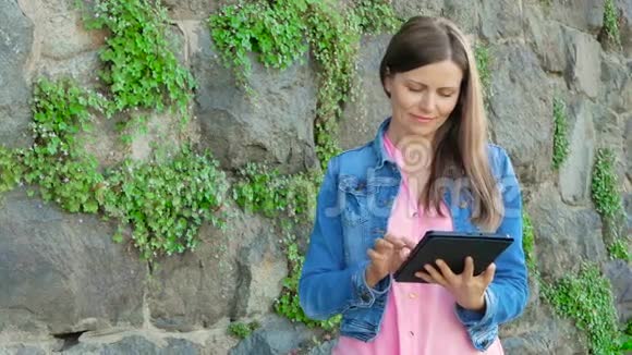 穿着棉夹克的漂亮女孩用平板电脑背景中野生石头的复古墙视频的预览图