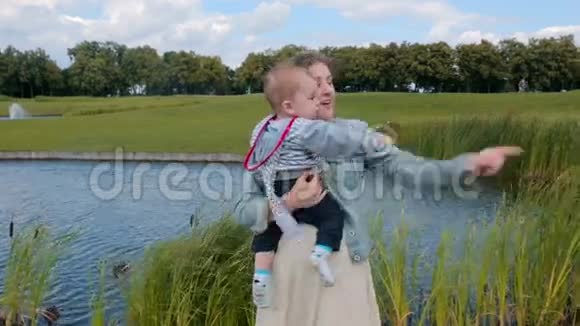 快乐的年轻母亲抱着她一岁的儿子在公园的大池塘里视频的预览图