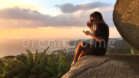 漂亮的女人坐在悬崖边用智能手机发短信泰国普吉岛高清慢动作惊人的视野视频的预览图