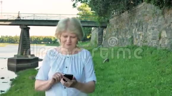 老年妇女沿着海滨散步使用智能手机背景中的野石桥和老墙视频的预览图