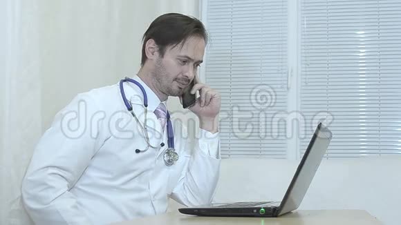 男医生在打电话医生在他的笔记本电脑办公室工作视频的预览图