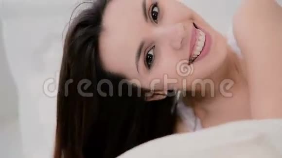 年轻的黑发女人在床上的肖像美丽的女孩躲在床单下对着镜头微笑视频的预览图