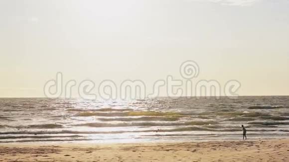 一个年轻女孩站在阳光明媚的沙滩上望着覆盖在她脚下的海浪视频的预览图