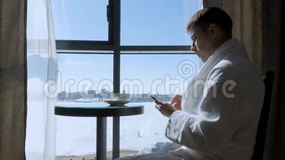 一个年轻有魅力的男人坐在靠窗的桌子旁喝茶喝咖啡在手机上写短信视频的预览图