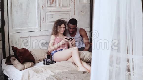 男人和女人穿着睡衣坐在床上多种族夫妇自拍照片使用智能手机和笑视频的预览图