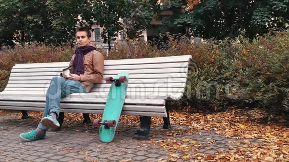 秋天公园里坐在长椅上的带着滑板的针织围巾的年轻人用纸杯喝咖啡视频的预览图