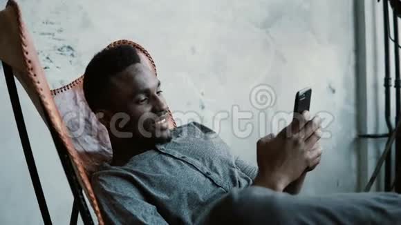 非洲男子坐在椅子上的肖像使用智能手机英俊的男性微笑看着手机里的照片视频的预览图