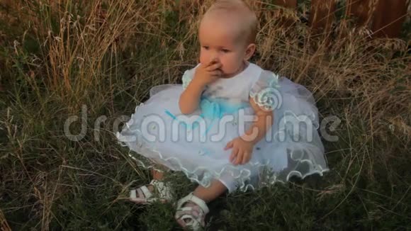 一个穿着白色连衣裙坐在高高的草地上的女婴她啃着杏子夏天日落视频的预览图