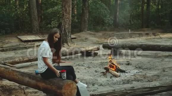 一个女孩坐在森林里看书女孩坐在火旁边的一根木头上看一本书特写视频的预览图