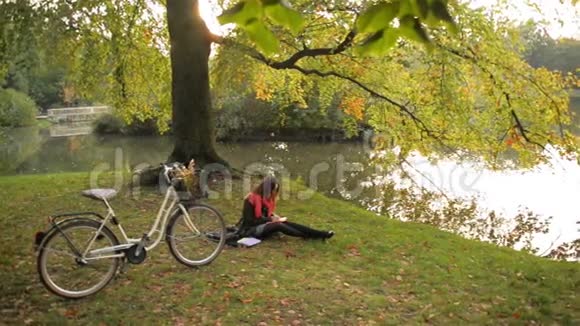 快乐的学生女孩在秋天的公共公园里放松在草地上看书背景中的自行车视频的预览图