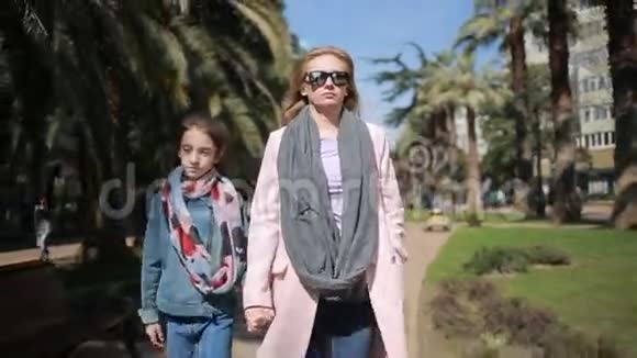 一个女人带着一个孩子母亲带着她十几岁的女儿在棕榈树的背景下沿着公园小巷走过视频的预览图