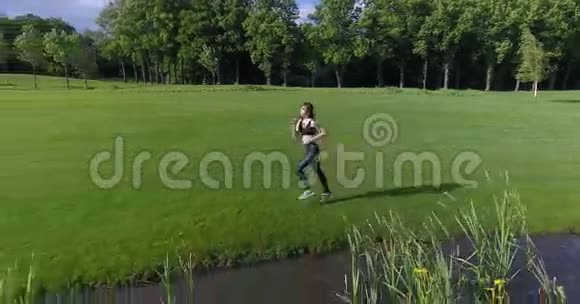 这位运动女子沿着河边的草地慢跑的水平视野移动的后视图视频的预览图