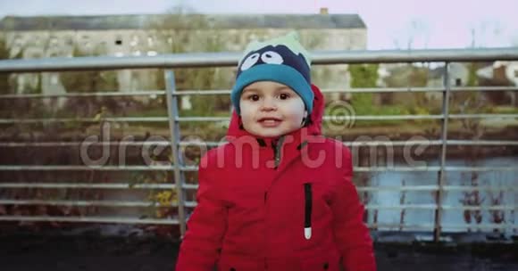 戴着红色夹克的可爱孩子在金属生锈栅栏前对着镜头微笑的肖像一幅美丽的肖像画视频的预览图