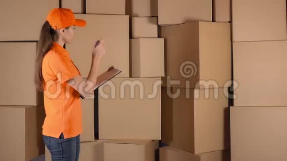 漂亮的女仓库工人穿着橙色制服计数物品并在棕色纸箱上做记录视频的预览图