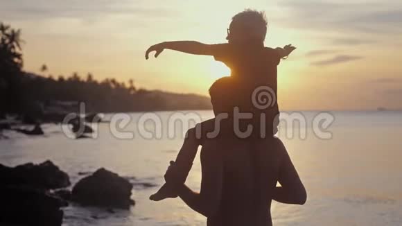 快乐的父亲和儿子戴着眼镜在热带海滩上玩耍男孩举起双手模仿美妙的日落飞行视频的预览图