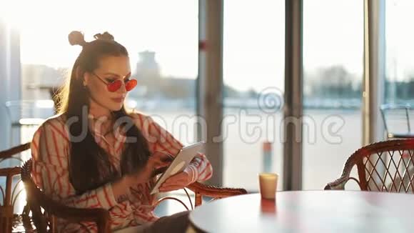 年轻迷人的女士戴着红色太阳镜坐在咖啡馆里的平板电脑机场或商店里的漂亮女孩视频的预览图