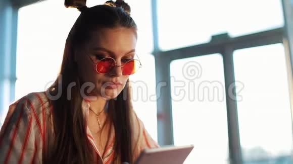 年轻迷人的女士戴着红色太阳镜坐在咖啡馆里的平板电脑机场或商店里的漂亮女孩视频的预览图