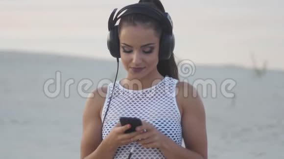 在海滩上使用手机智能手机的女人穿着耳机听音乐或者在比基尼上用智能手机视频的预览图