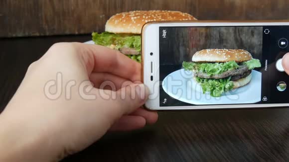女性手在智能手机上制作一个漂亮的多汁汉堡在时尚的木制上放生菜叶视频的预览图