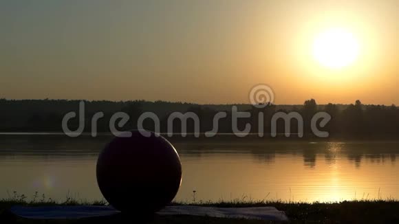 粉红色的健身球躺在太阳落山时湖岸的瑜伽垫上视频的预览图