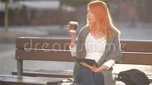红发女孩坐在街上的长凳上喝咖啡年轻女子在阳光明媚的日子里用平板电脑城市视频的预览图