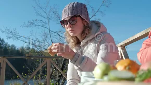 年轻女子穿着暖和的衣服吃着汉堡狗在附近玩耍在河边的木桥上野餐周末视频的预览图