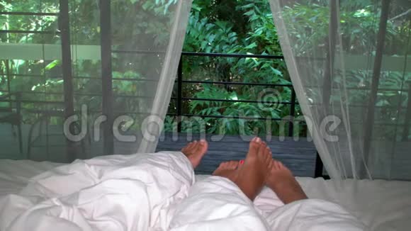 浪漫的情侣睡在床上白色的窗帘和双脚朝窗外敞开着茂密的热带绿色植物视频的预览图